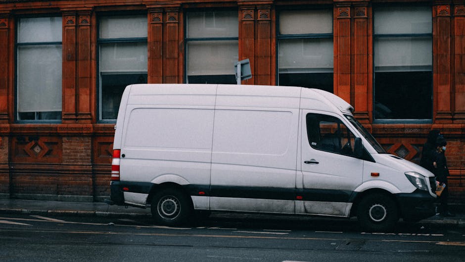 A white commercial van belonging to Removal Van Brixton is parked on a city street next to a multi-storey building. The van's rear doors are wide open, revealing a load of various packing materials an