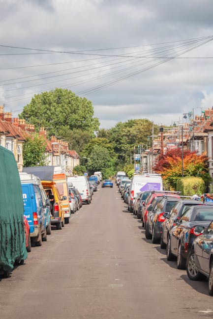 A daytime street scene on Coldharbour Lane in Brixton with a large mature tree and a small brick building in the background. Several pedestrians are walking along the pavement, some near bicycles parked against the building's wall. A black van is parked on the right side of the image, with its rear doors open, suggesting a loading or unloading process. The street has yellow cross-hatched markings indicating a no-parking zone, and a 20 mph speed limit sign is visible on a lamppost. The sky is partly cloudy with patches of blue, and the area appears to be part of a community setting suitable for local house removals, with Removal Van Brixton potentially involved in furniture transport and home relocation activities within the neighbourhood.