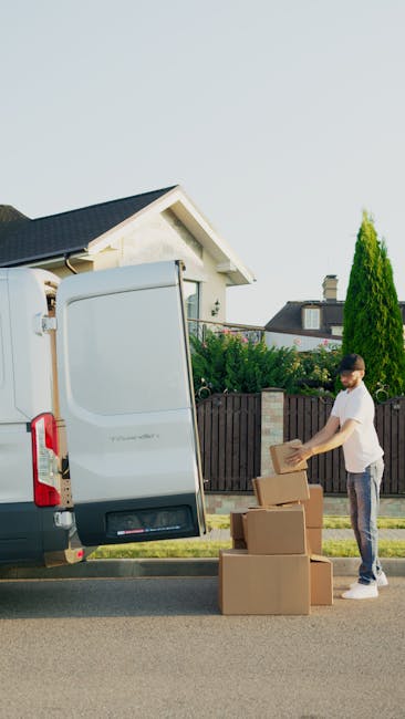 A man sitting on a wooden chair outside a white removal van during a house relocation, with the van's open side door revealing empty wooden packing crates and cardboard boxes. The man is wearing glass